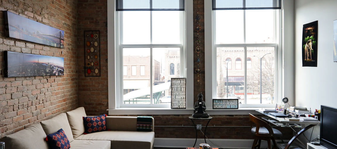 Loft apartment with exposed brick, big windows, beige sectional sofa, patterned pillows, wall art, Buddha statue, and corner desk.