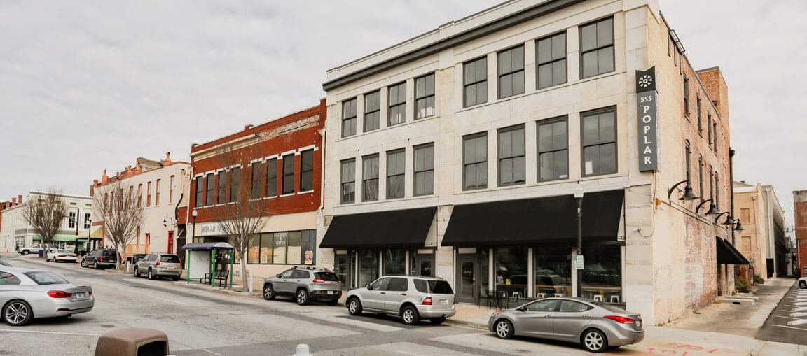 A city street with parked cars in front of a three-story building with large windows, black awnings, and a vertical 555 POPLAR sign.