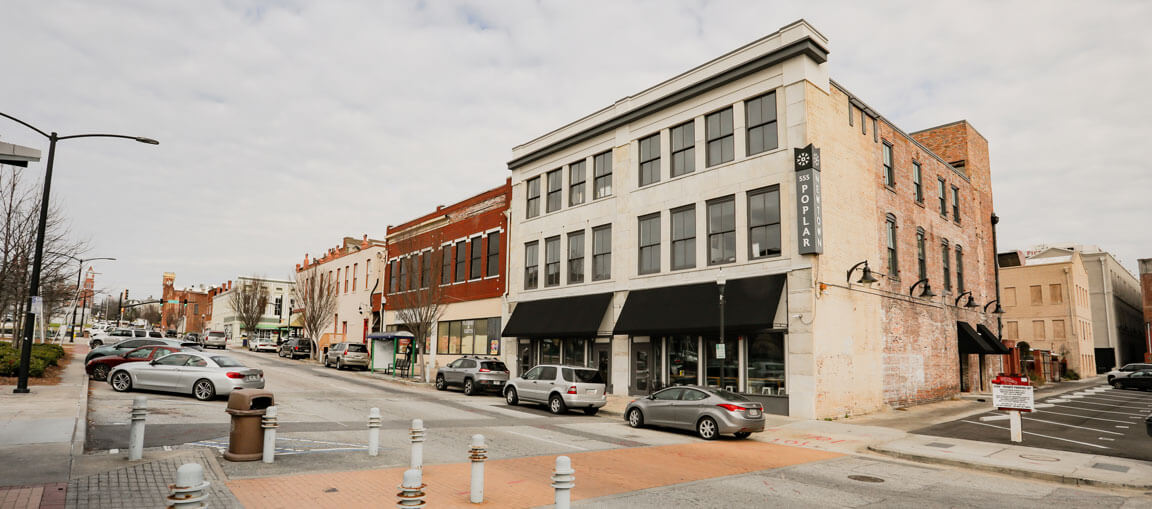 A city street with several parked cars and brick buildings, including a corner building with black awnings and a Poplar sign. The sky is overcast and a few pedestrians are visible in the distance.