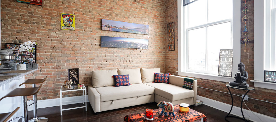Cozy living room with brick walls, beige sectional, colorful pillows, wall art, large window, barstools, Buddha statue, eclectic decor.