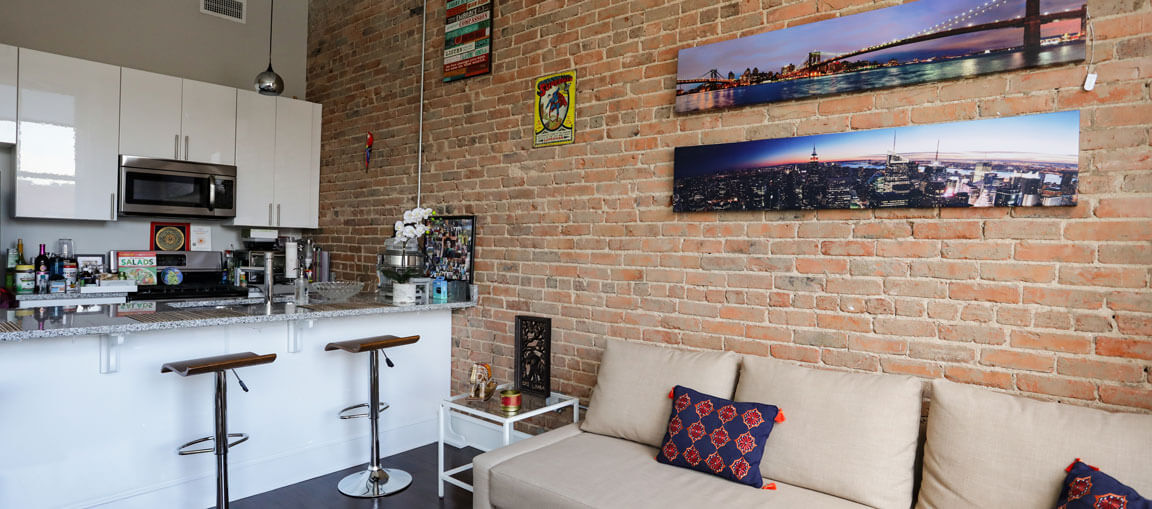 Modern loft apartment with exposed brick walls, beige sofa with patterned pillows, small kitchen with barstools, cityscape photos above.