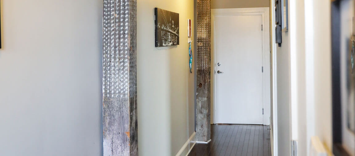 A well-lit hallway features light walls, dark wood floors, metal panels, wall art, and a closed white door at the end. Modern decor.