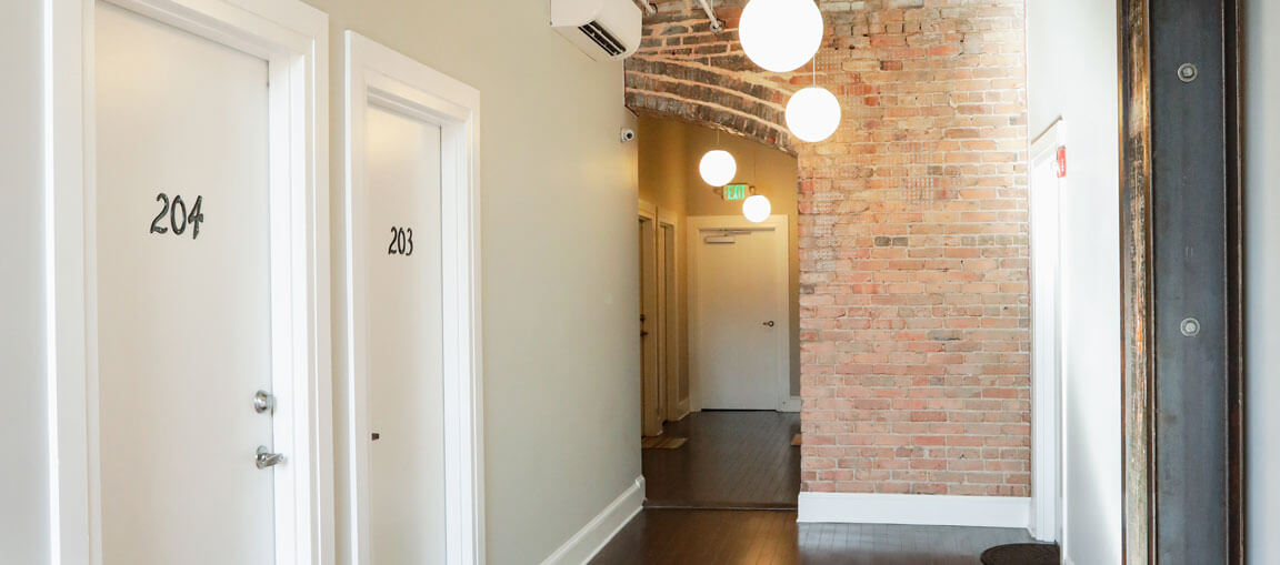 A hallway with white doors 204 and 203, exposed brick wall, round hanging lights, white walls, dark wood floor, more doors ahead.