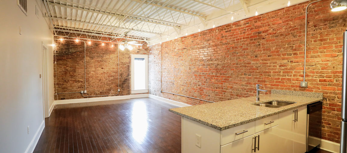 Open loft apartment with exposed brick walls, dark wood floors, white kitchen cabinets, granite countertop, beams, and string lights.