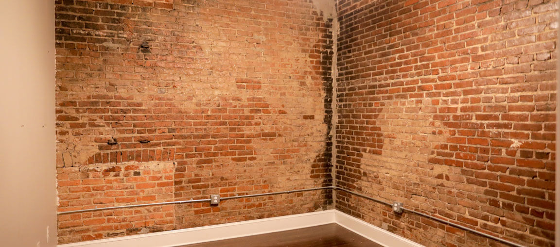 Empty room with exposed red brick walls, moisture-darkened spots, white baseboards, dark wood floor, and metal conduit along walls.
