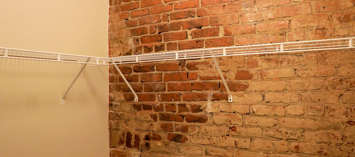 White metal wire shelves on a corner wall, one side beige-painted, the other exposed red brick; shelves empty, angled brackets.