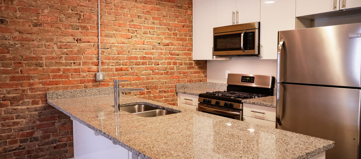 Modern kitchen featuring exposed brick wall, granite counters, stainless steel appliances, and a kitchen island with sleek faucet sink.