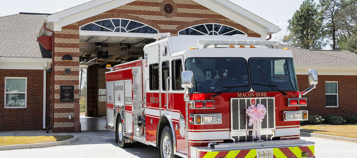 A red and white fire truck decorated with a pink bow is parked outside a brick fire station with an arched entrance on a sunny day.