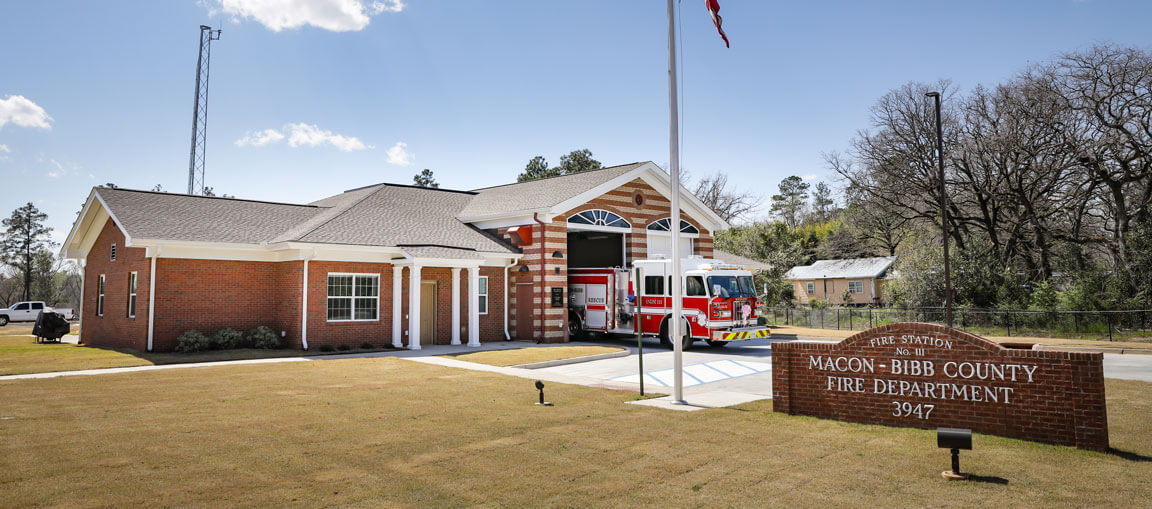 A red and white fire truck outside the Macon-Bibb County Fire Department with a brick sign, green lawn, trees, and clear sky.