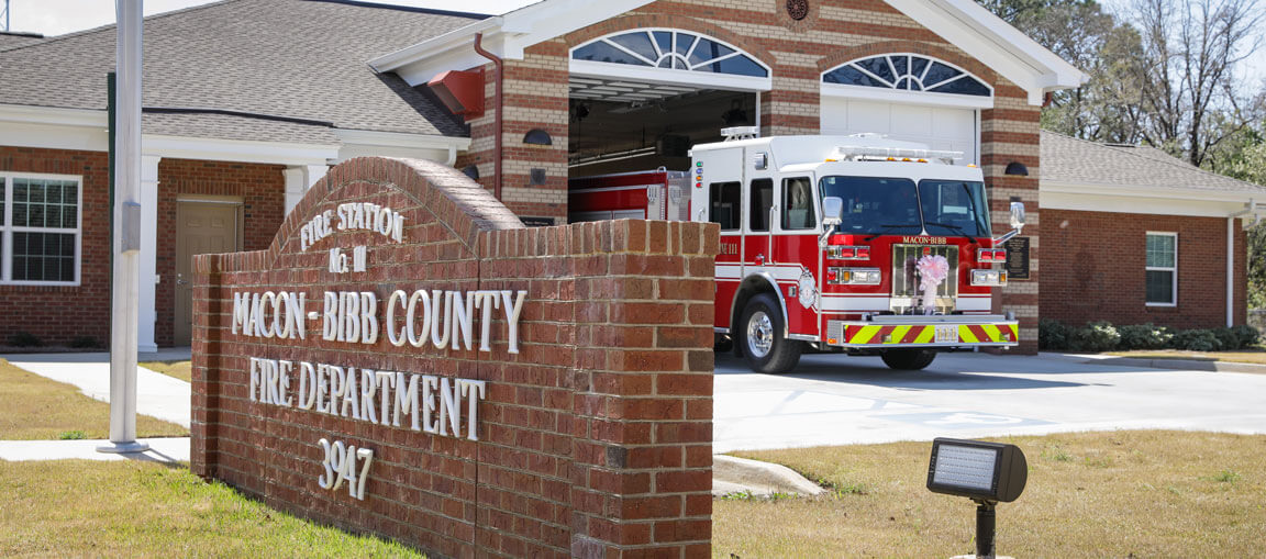 A red and white fire truck is parked outside a brick fire station with a sign reading Macon-Bibb County Fire Department 9047 in front of the building.