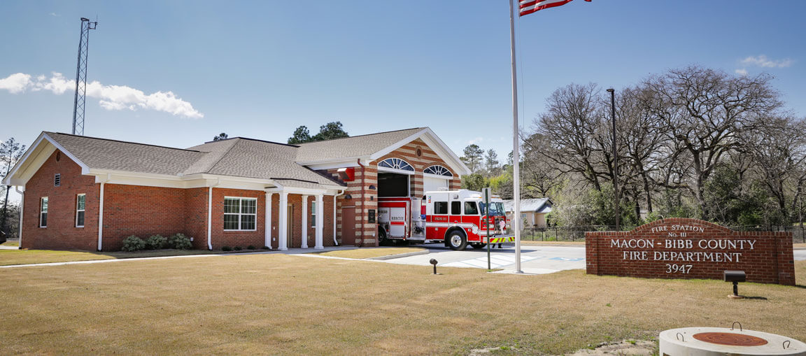A red fire truck is parked outside the Macon-Bibb County Fire Department, with a U.S. flag and brown sign in front of the brick station.