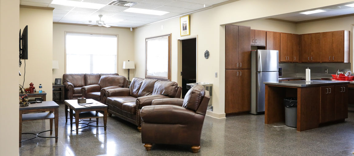 Spacious living room and kitchen with brown leather sofas, coffee table, wooden cabinets, and bright natural light from large windows.