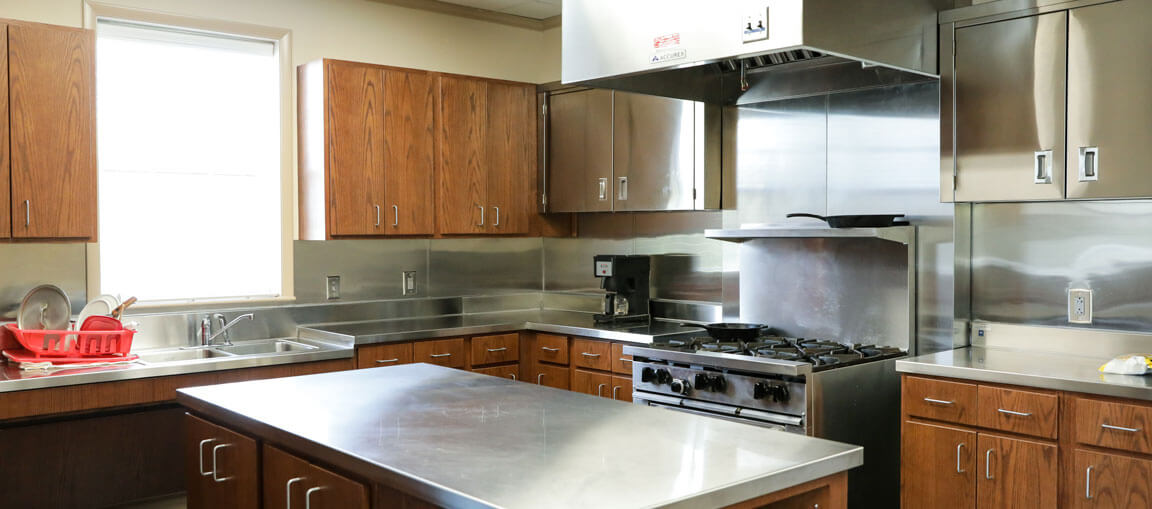 Commercial kitchen featuring wooden cabinets, a stainless steel island, gas stove, metal backsplash, and a red dish rack near the window sink.