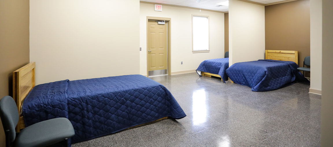 A simple room with two single beds in blue quilts, gray chairs beside each, beige walls, a door, window light, and gray floor.