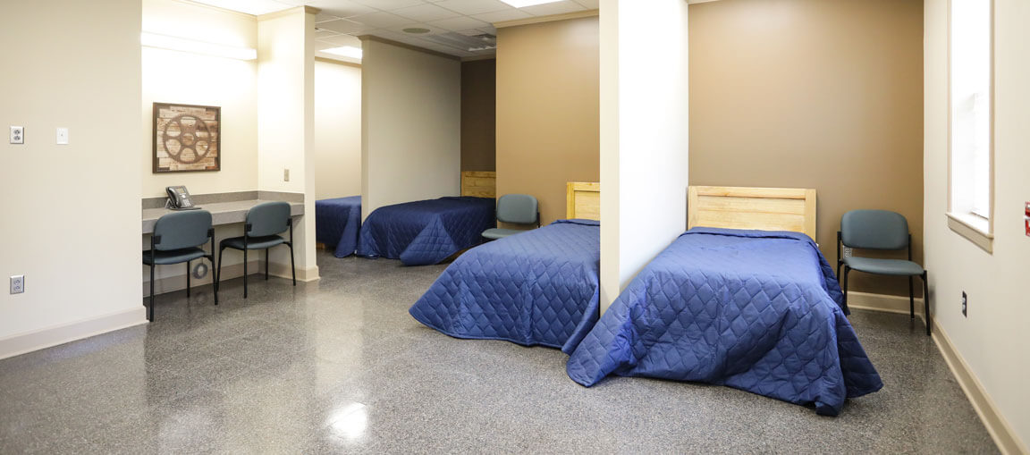 A clean, sparsely furnished room with three single beds in blue quilts, wooden headboards, gray chairs, desk, and neutral walls.