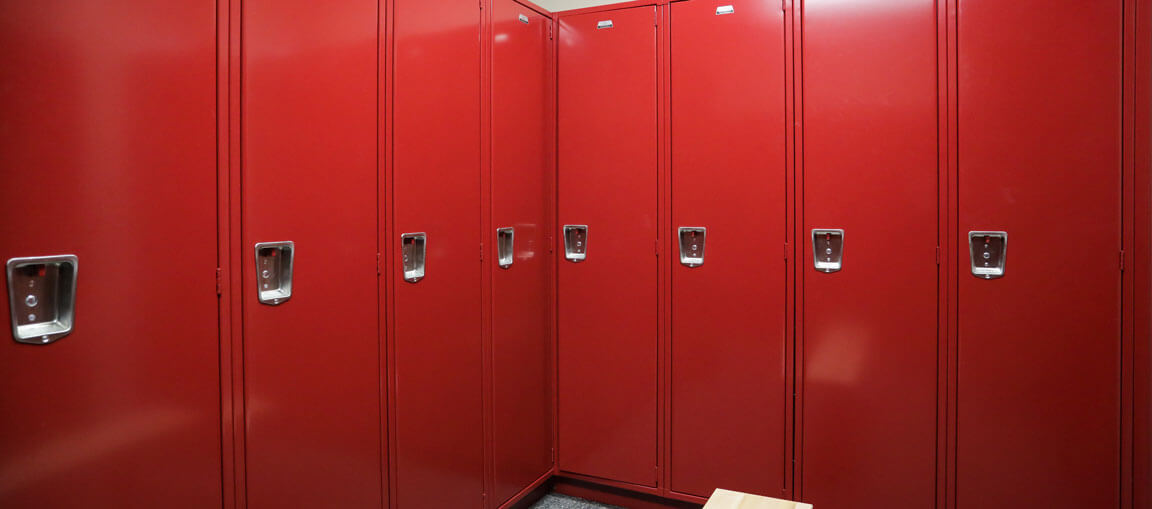 A row of tall red metal lockers with silver handles lines a corner. The dark floor and part of a wooden bench are also visible.