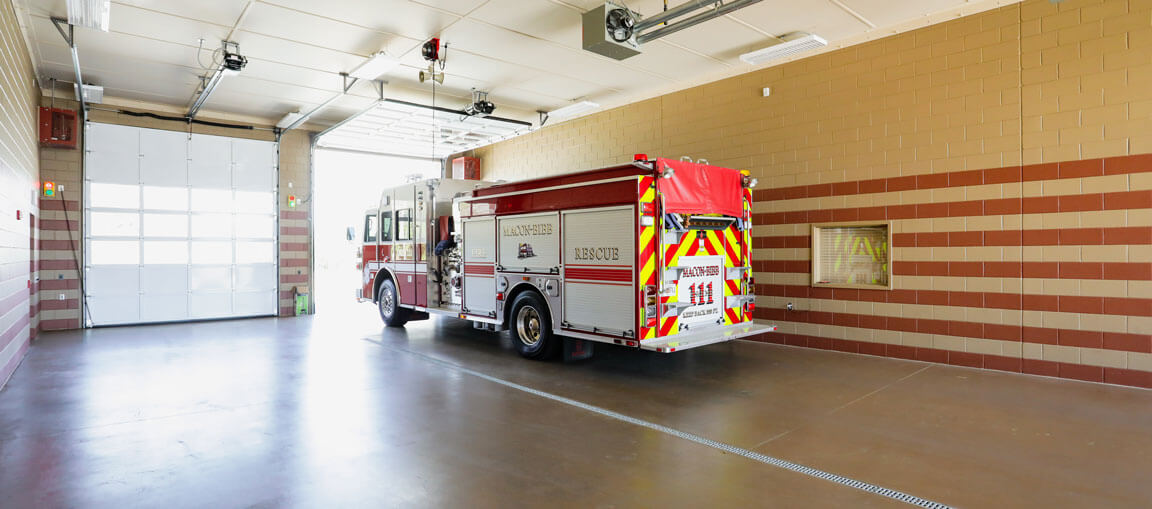 A fire truck is parked inside a fire station garage, rear toward the open door. Beige and brick walls, polished concrete floor.