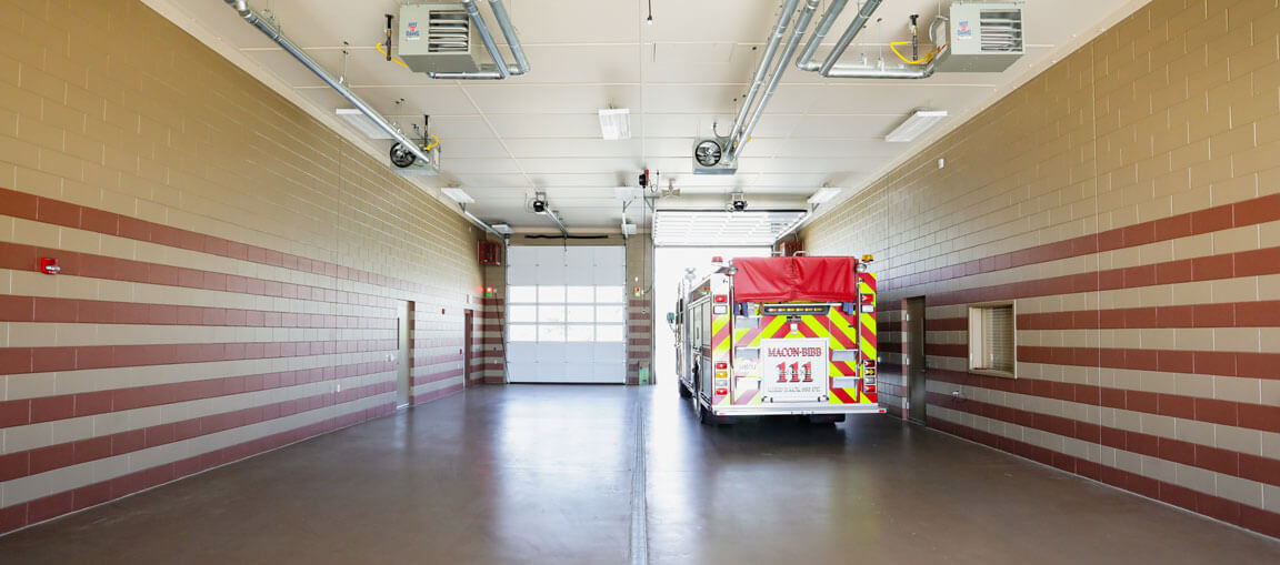 A fire truck is parked in a clean, empty fire station garage with tan and brown striped walls, overhead lights, and an open door.
