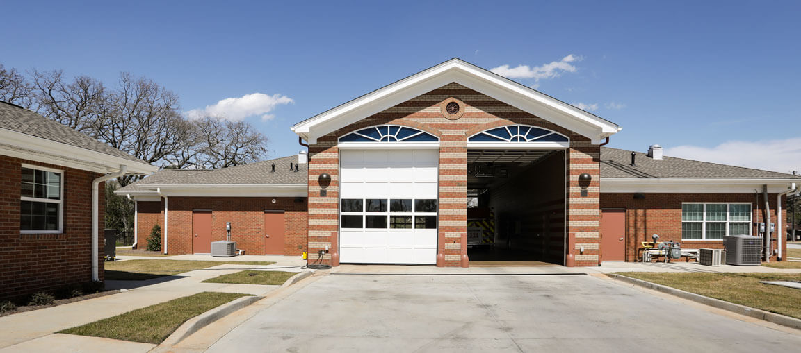 Modern brick fire station with large white garage door open to show a fire truck, flanked by wings and a paved driveway, sunny sky above.