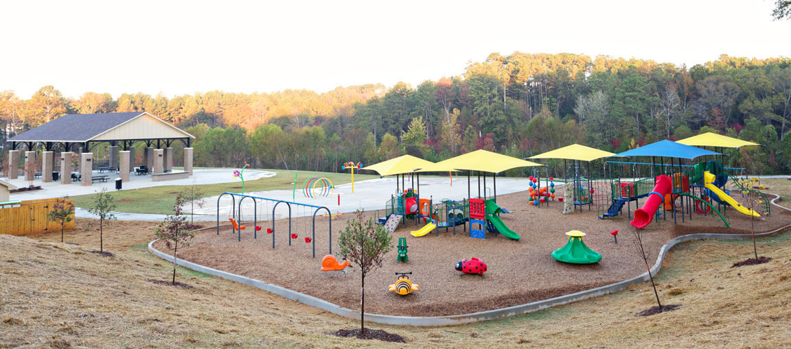 A large playground outdoors with slides, climbing frames, swings, bright shade covers, trees, grass, and a pavilion in the background.