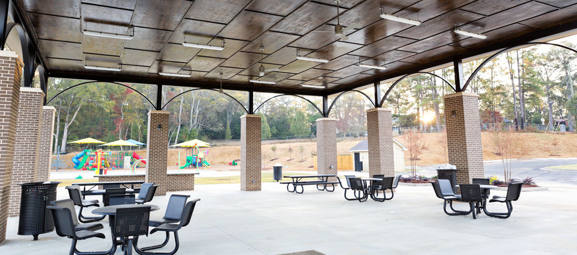 Covered outdoor patio with round tables, chairs, brick columns, and lights. Playground with slides and greenery visible in back.