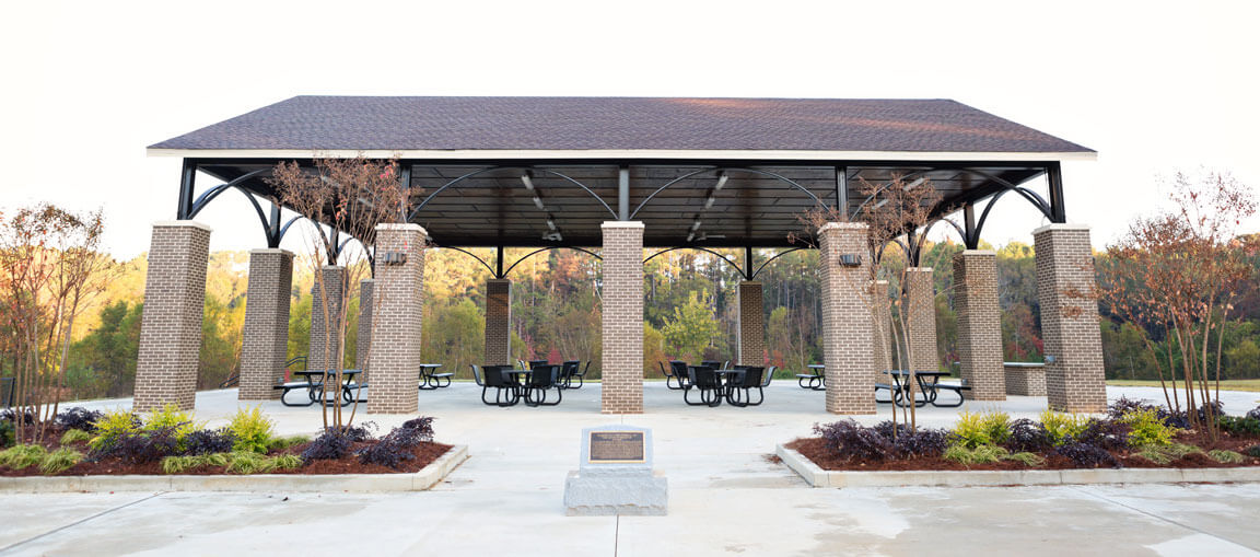 Open-air pavilion with brown roof on brick columns, several round picnic tables, landscaped trees and shrubs in front, trees behind.