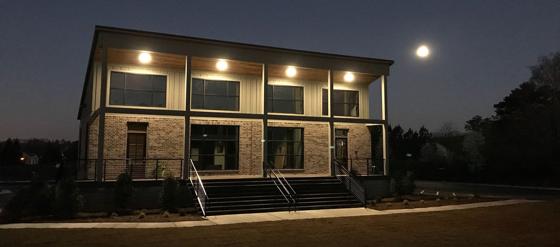 A two-story brick building with large windows glows at night. A staircase, small shrubs, sidewalk, and full moon complete the scene.