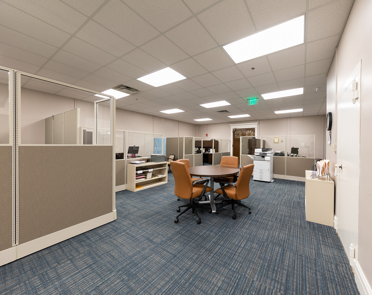Modern office with blue carpet, beige cubicles, round table and four brown chairs, computers on desks, printer in back corner.