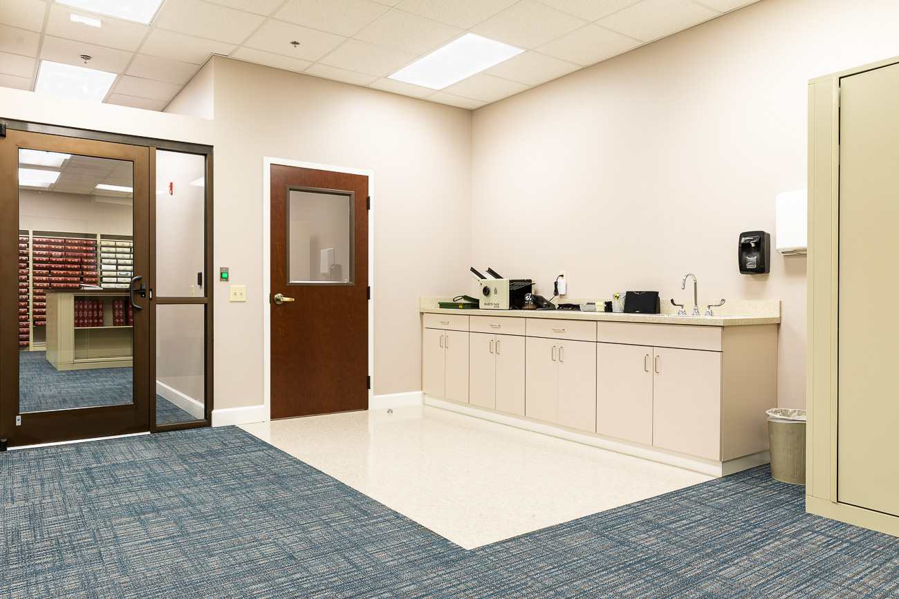 Clean, modern office with blue carpet, beige cabinets, sink, supplies on counter, brown door, glass entrance, filing shelves.