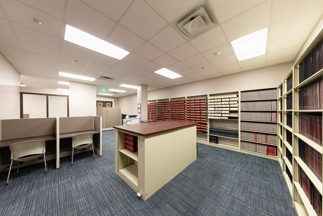 A spacious, well-lit library with shelves of books and files, a large table, study cubicles, blue carpet, and cream walls.