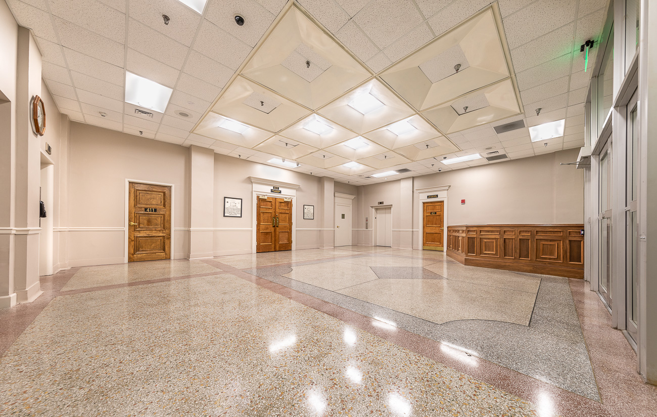 A spacious empty hallway with polished floors, wooden doors, bright tile ceiling lights, wood panel walls, and a far-end elevator.