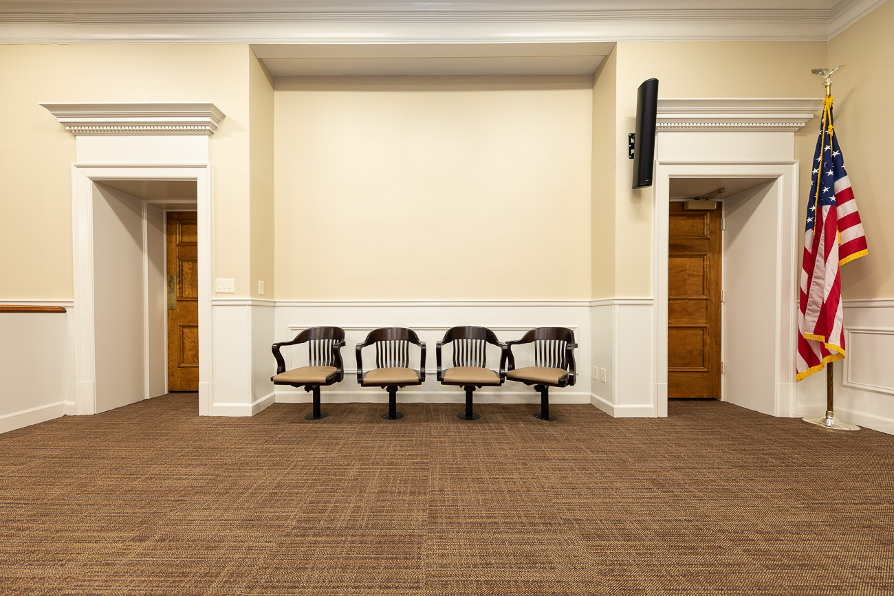 Four empty chairs in a row against a beige wall, brown carpeted floor, two doors on each side, American flag in the corner.