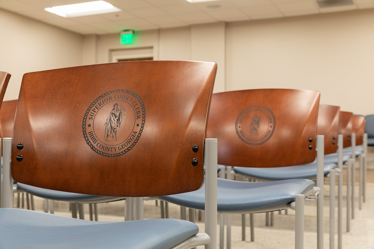 Rows of wooden chairs with metal frames and blue seats in a courtroom, each chair back engraved with the Bibb County, Georgia seal.