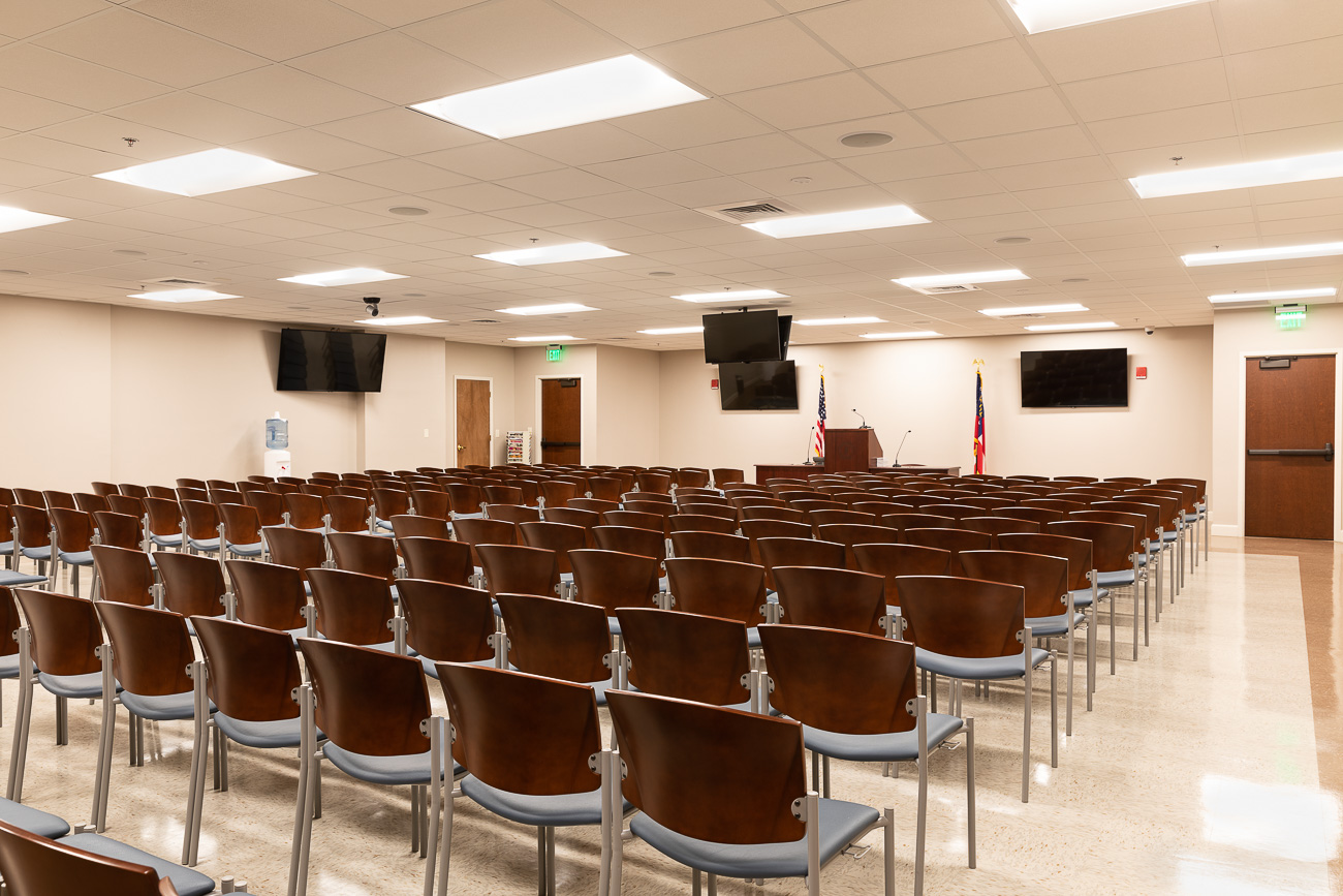 A well-lit conference room with beige walls, rows of empty brown chairs, a podium, screens, and American and state flags in front.