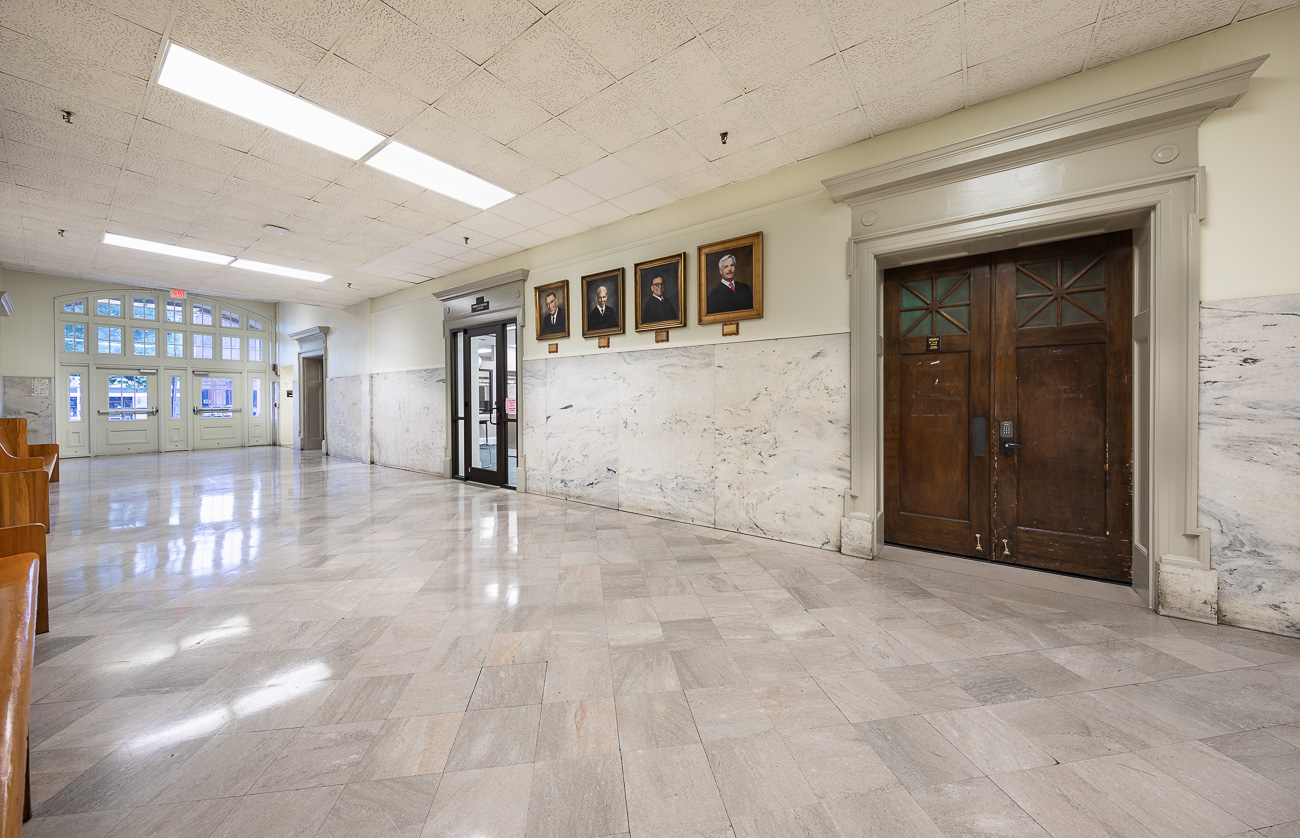 Bright, empty hallway with marble floors and walls, wooden doors and portraits on the right, large windows and benches on the left.