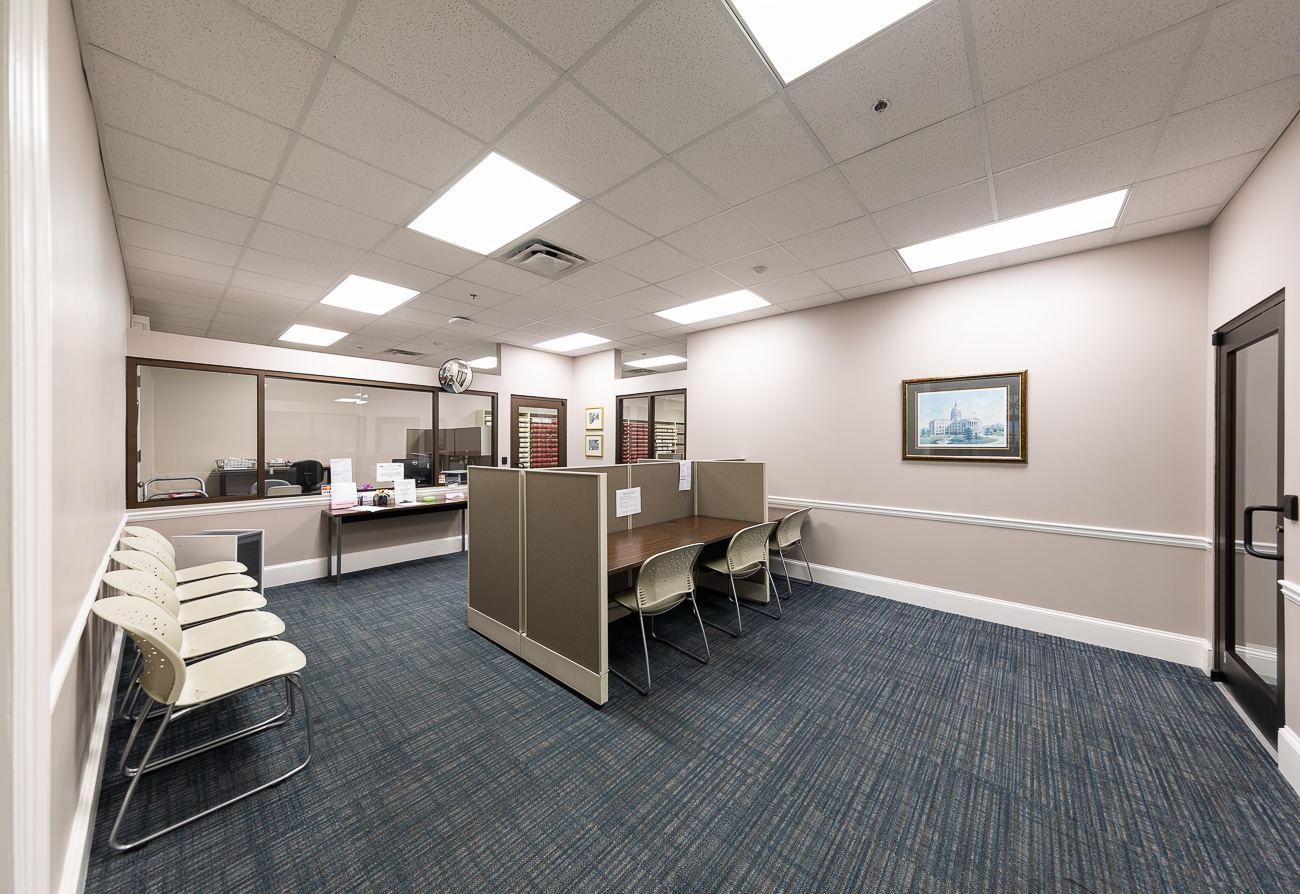 Clean modern office with cubicles, blue carpet, beige walls, chairs by the wall, overhead lights, and a glass partition to an office.