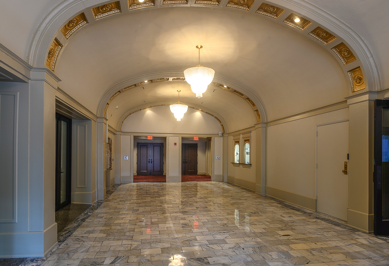 A wide, elegant hallway with marble flooring, gold-accented arched ceiling, and chandeliers. Two elevator doors are at the end.