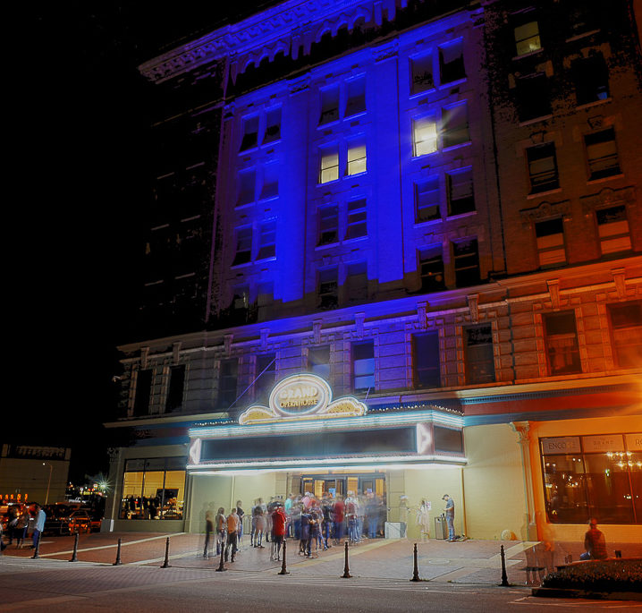 Nighttime scene of a theater entrance, crowd outside. Blue and orange lights illuminate the building’s details against a dark sky.