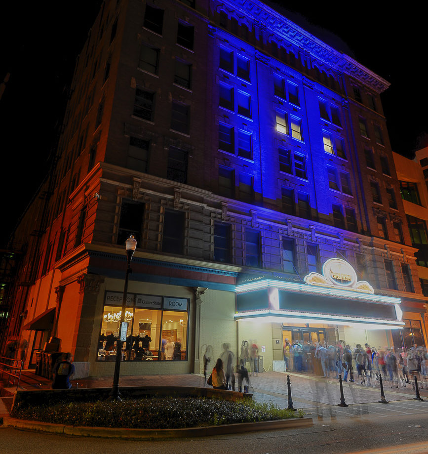 A large theater lit by blue and yellow lights at night, people gather near the entrance with motion blur, creating a lively urban feel.