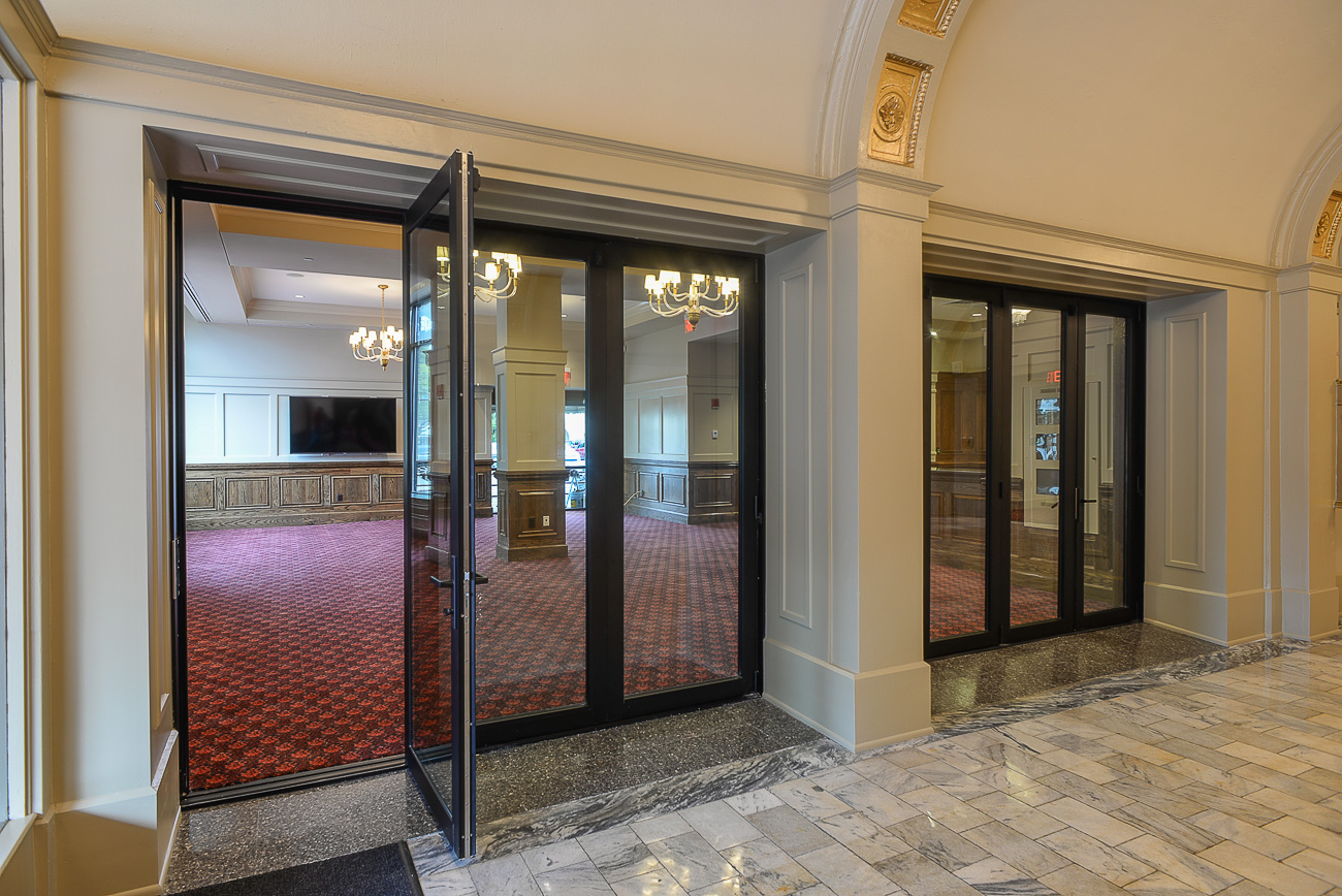 Glass double doors lead to a grand room with red patterned carpet, chandeliers, wooden paneling, marble entry floor, and arches.