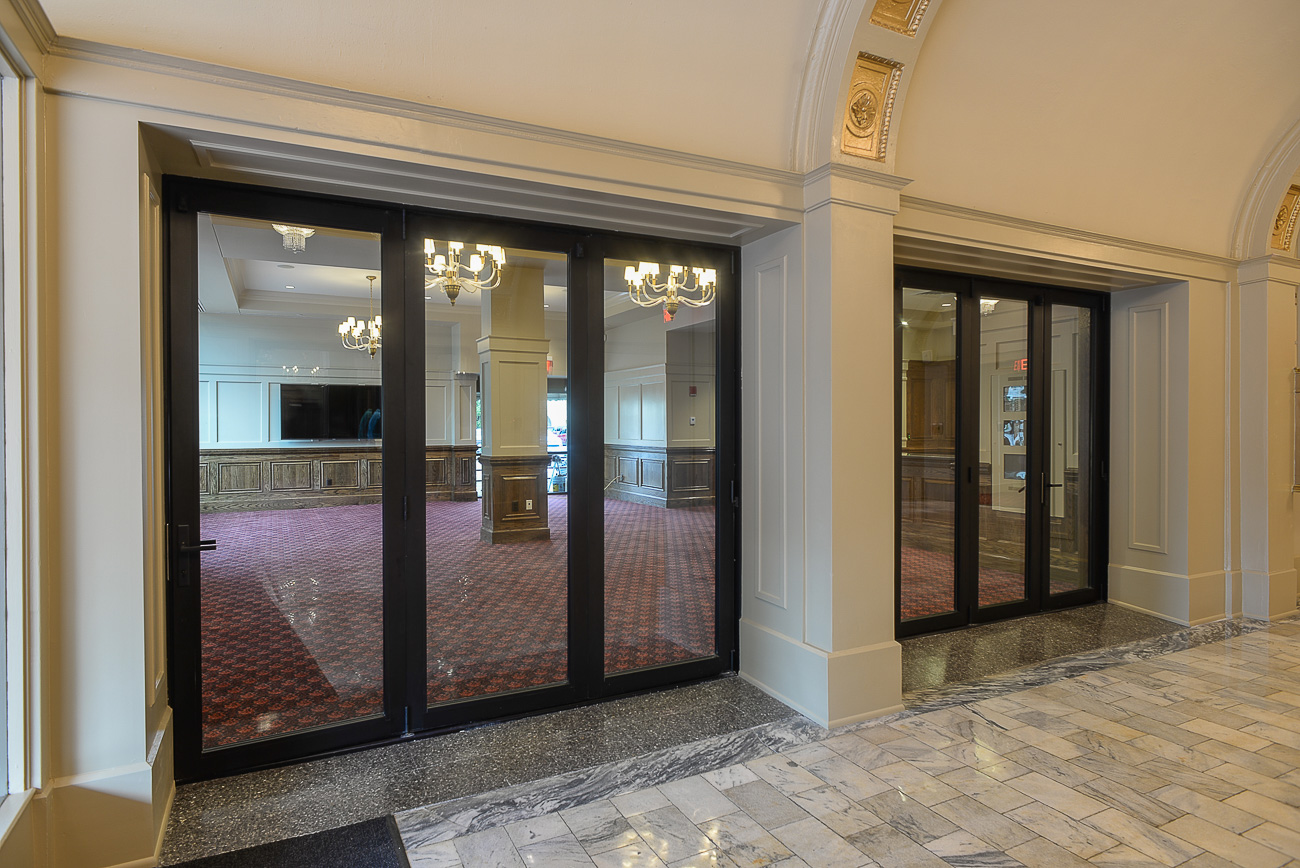Glass double doors with black frames open to an elegant interior featuring chandeliers, red carpet, marble floors, and ornate molding.