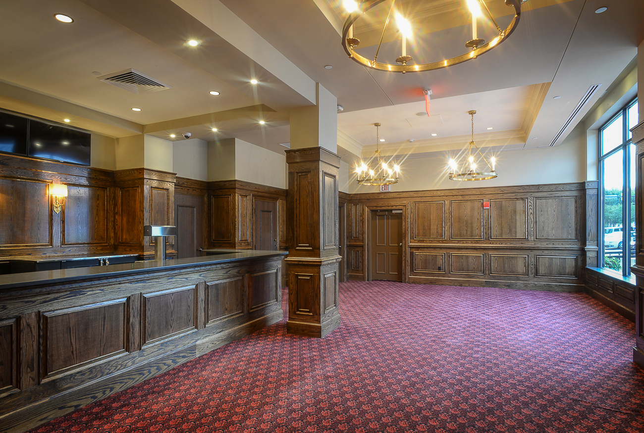 Spacious, elegant room with wood-paneled walls, red patterned carpet, large windows, chandeliers, and a long wooden counter.