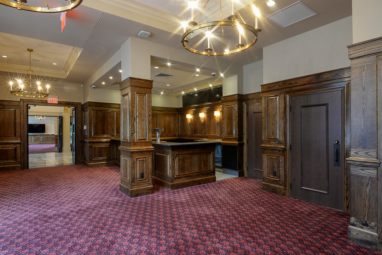 A spacious lobby with dark wood paneling, ornate crown molding, red patterned carpet, modern chandeliers, and multiple doorways.