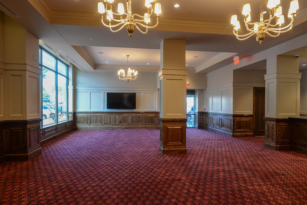 Spacious empty room with red patterned carpet, beige walls, wood trim, columns, chandeliers, large windows, TV, and natural light.