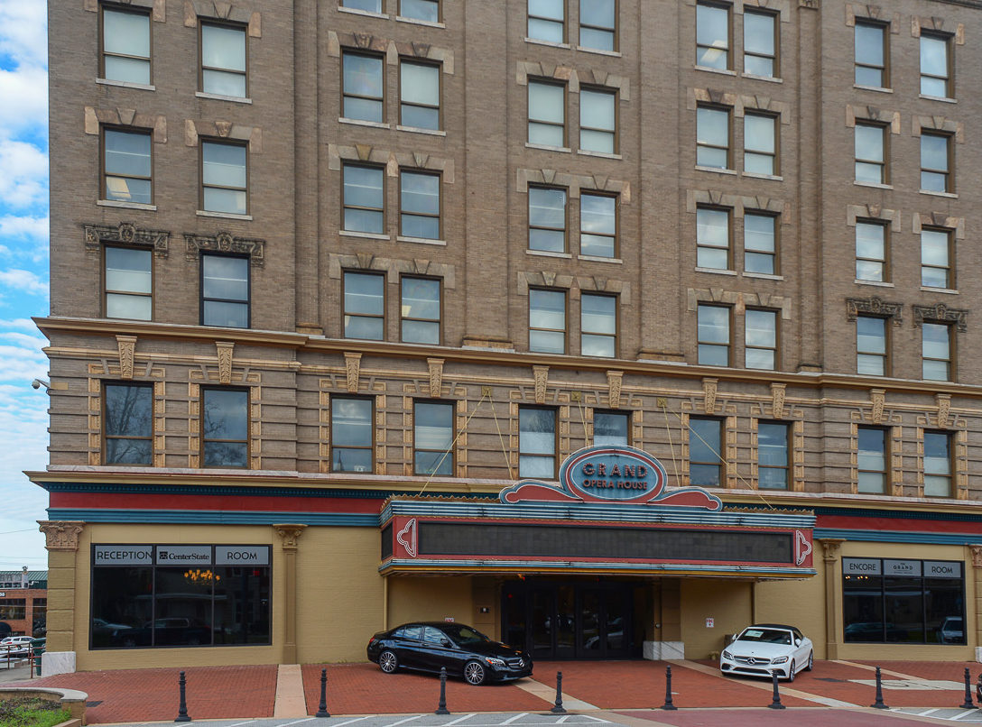 A historic multi-story brick building with a large marquee reading Grand Opera House above the entrance, two cars parked in front.