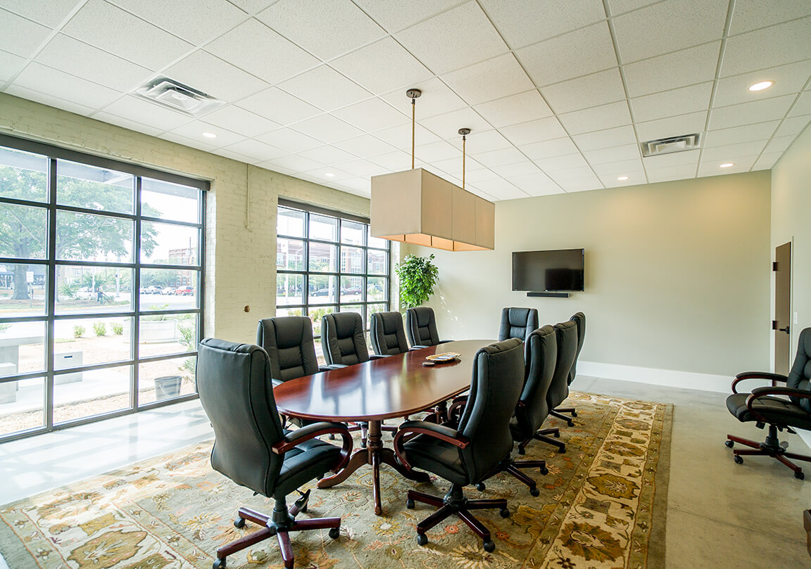 large conference room with a long wooden table, 10 chairs, all on a rug, with a tv on the wall, with 2 garage door style windows