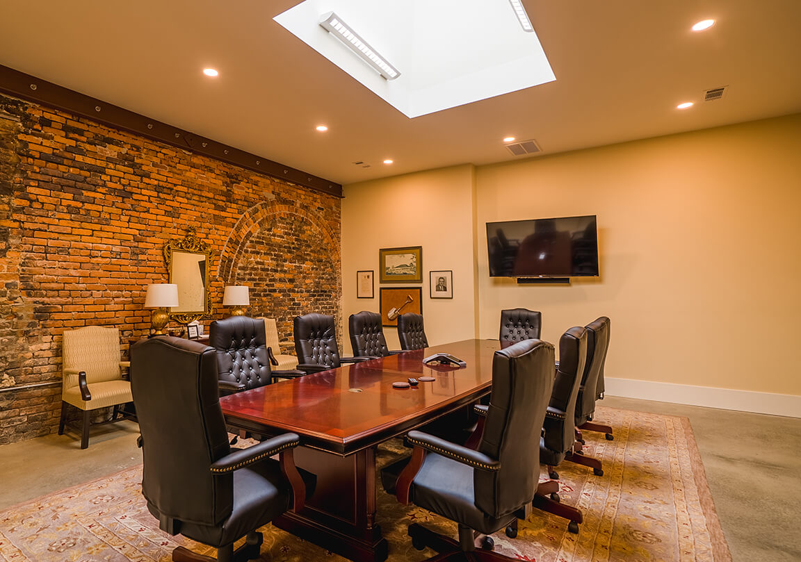 a large wooden table in a conference room that is surrounded by 10 chairs, an exposed brick wall with 2 additional chairs and a side table, and a tv. There is also a skylight bringing in natural light.