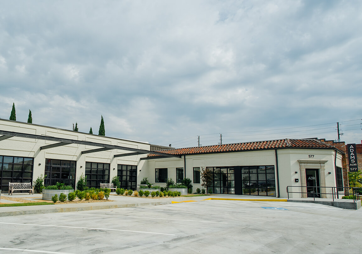 outside of the Adams ADR building, white brick, a lot of dark garage door style windows, and a patio area with benches, tables, and plants, ramped sidewalk to the front door