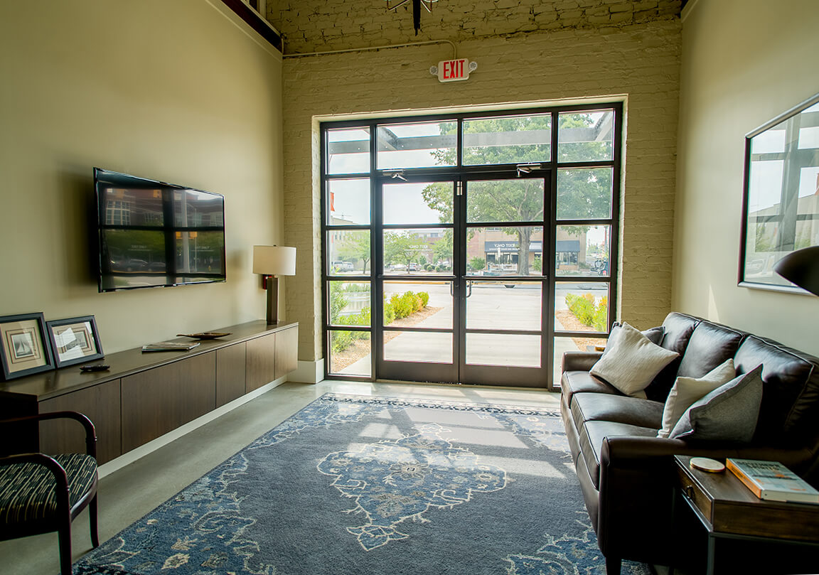 a waiting room with a comfortable couch with a window behind it, rug on the floor, side tables, tv
