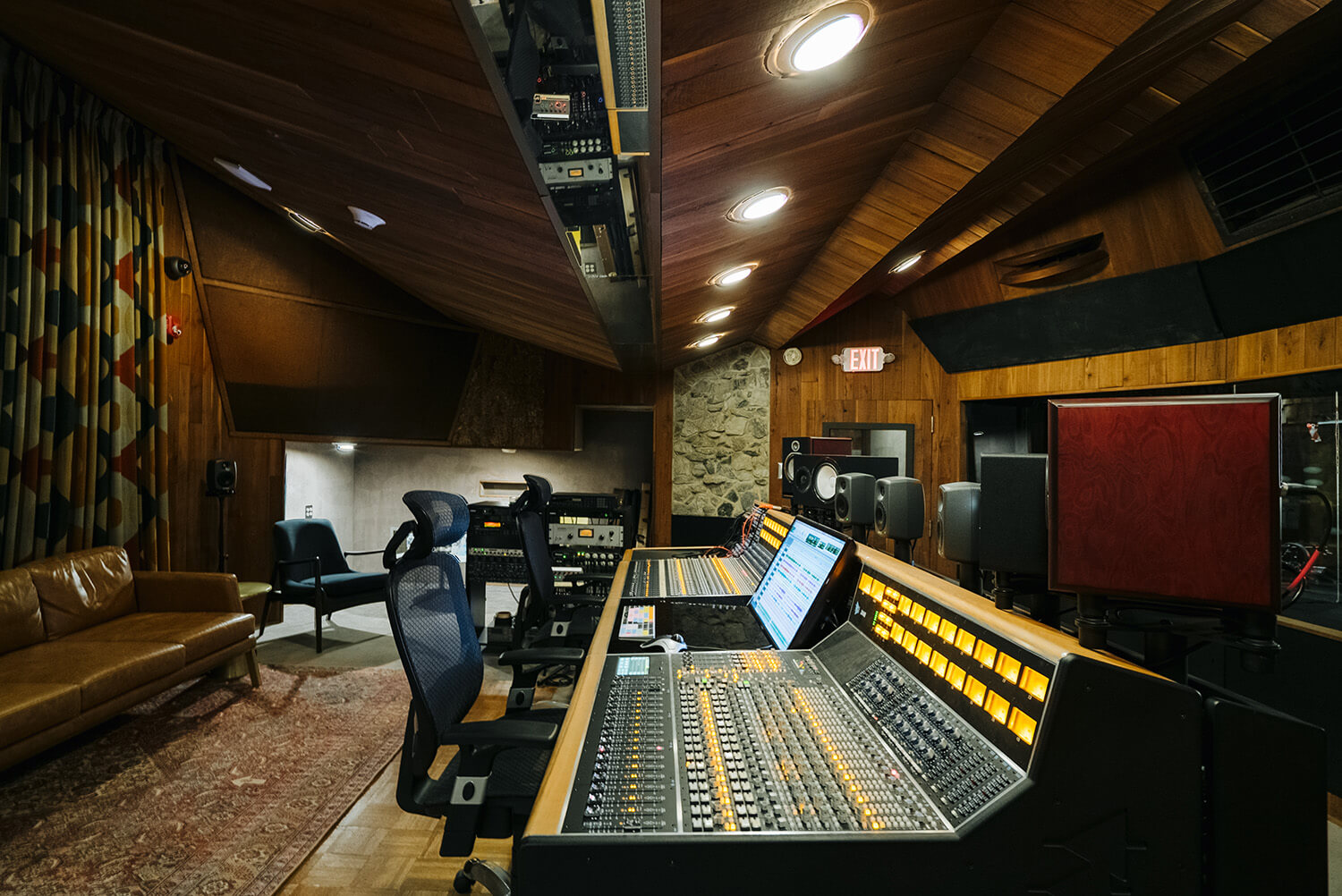Professional recording studio with large mixing console, monitors, chairs, brown leather sofa, patterned curtains, and wood-paneled walls.